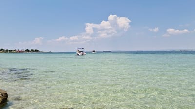 This media file depicts a serene seascape with clear, shallow turquoise water dominating the foreground. Sunlight glints off the rippling surface, revealing the sandy seabed and scattered rocks beneath. In the middle ground, two small motorboats are visible on the water. One boat, closer to the viewer, has a canopy and several people aboard, suggesting a leisure activity. The second boat, further out, also appears to be carrying people. In the distance, a large cargo ship is present on the horizon, indicating shipping activity.
The background features a coastline with a line of trees and buildings, suggesting a populated area. The sky above is a vibrant blue, punctuated by scattered white clouds, with one particularly large, fluffy cloud resembling a heart-shaped formation positioned above the boats. The overall impression is one of a bright, sunny day, likely during the daytime, suitable for water recreation. The clear water and tranquil scene evoke a sense of peace and relaxation. The location context provided, Taranto, Italy, suggests a Mediterranean setting. There is no visible text in the image.