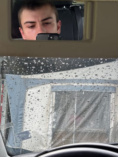 A person is taking a selfie in a car mirror. The car window next to the mirror is covered in raindrops. Outside the window, the scene is blurry, possibly an urban environment. The location is Dabovets, Bulgaria.