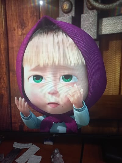 The image displays a close-up of a cartoon character, Masha, from the animated series "Masha and the Bear." Masha is depicted with her signature purple headscarf and blue outfit. She has large green eyes and her right hand is raised to her face, appearing to be touching her cheek or wiping away a tear, suggesting a moment of sadness or contemplation. The background is dimly lit and appears to be indoors, with wooden paneling on the left and a textured surface on the right.
Below the screen, there are several blister packs of medication and a small blue box with text visible, suggesting the image was taken in a home environment. The presence of the LG logo on the bottom bezel of the screen indicates it is displayed on a television. The image is taken from a slightly elevated angle, looking down at the screen. The location of Calarașovca, Moldova is not visually represented in the image itself.