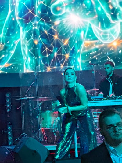 The image depicts a dimly lit stage with a woman in a shimmering green outfit in the foreground, seemingly performing or posing. Behind her, a man with reddish hair is operating a DJ setup. A drum set is visible further back, illuminated by colorful stage lights. A large, abstract, glowing digital art piece forms the backdrop, featuring interconnected geometric shapes and bright lights. The overall scene suggests a concert or a performance event taking place indoors. The exact location in Akhalubani, Georgia, cannot be determined from the visual information provided in the image.