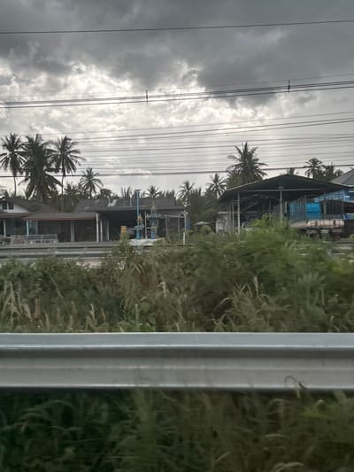 The image depicts a view of Ban Khlong Chai Tong, Thailand, dominated by a stormy sky and a line of utility poles with numerous wires. On the right side, a silver metal barrier is prominent, with lush green foliage behind it. The scene is composed of a mix of natural and man-made elements.

The background features several buildings, some appearing to be workshops or storage facilities with open sides and corrugated metal roofs. Palm trees are scattered throughout the landscape, their fronds silhouetted against the overcast sky, suggesting a tropical or subtropical setting. The weather appears inclement, with heavy, dark clouds filling the sky and a diffused light indicating a possibly stormy or overcast day.

There are no visible people in the image. The focus is on the infrastructure, vegetation, and the dramatic sky. The presence of utility poles and wires suggests an inhabited area, likely a roadside or a village. The overall mood is somewhat somber due to the weather conditions. No legible text is visible in the image. The time of day is indeterminate due to the lack of strong shadows and the overcast conditions.