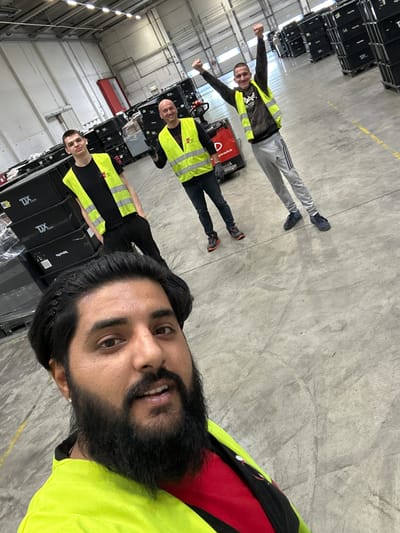 In a warehouse environment in Schweitenkirchen, Germany, three men are visible. In the foreground, a man with dark hair and a beard wearing a bright yellow safety vest over a red shirt is taking a selfie. Behind him, two other men, also wearing yellow safety vests, are in the background. One is looking at the camera, while the other has his arms raised in a celebratory gesture. Stacks of black storage containers with "TDX EUROPE" branding are visible in the background, suggesting a logistics or distribution facility. A red pallet jack is also present. The floor is concrete, and overhead lighting illuminates the space.