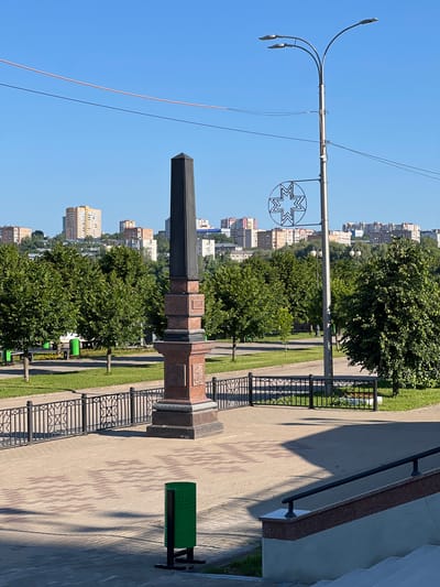 In Sovetsko-Nikol'skoe, Russia, a monument stands tall in a city park. The monument, dark and rectangular at its apex, rests on a multi-layered, granite base. Inscriptions and ornate carvings adorn the base. The park itself is meticulously maintained with green lawns and mature trees. A black metal fence delineates the park area. Paved pathways wind through the park, leading to benches and green trash receptacles. In the background, a modern street lamp with multiple lights soars towards the clear blue sky. A decorative, star-shaped metal ornament hangs from the lamp post. Beyond the park, a cityscape unfolds with a mix of apartment buildings and older structures, nestled against a backdrop of rolling hills. The sunlight casts sharp shadows across the scene, indicating a bright, clear day. There are no people visible in the frame.