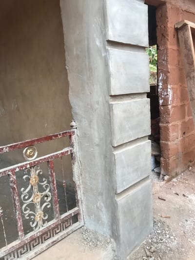 This daytime photograph, captured in Ogodo, Nigeria, depicts an outdoor construction or renovation area. A central, light grey concrete pillar, recently plastered, features five stacked rectangular relief elements. To the left, a section of a reddish-brown wrought iron gate or railing is present, exhibiting white and gold scrollwork and a Greek key motif along its base. This gate segment connects to a light brown plastered wall in the background. To the right of the concrete pillar, an unlit opening or doorway reveals indistinct green foliage beyond. A red-brown brick wall, rough and unfinished, forms the right boundary of this opening, with two wooden planks leaning against it. The ground is unpaved, consisting of dirt and gravel, with scattered small concrete fragments and debris at the base of the pillar and brick wall. No individuals or active events are observed.