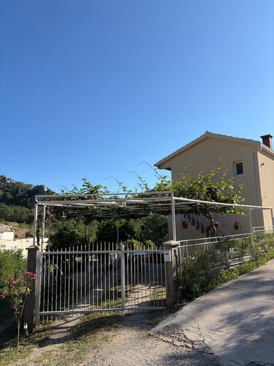 The image shows a view from an outdoor perspective, with a clear blue sky dominating the left side. A house with a cream-colored facade and a terracotta tiled roof is visible on the right, partially obscured by a white metal pergola. The pergola is covered with lush green vines, some of which have small red flowers. Potted plants hang from the pergola. A metal gate, also white, stands in front of the pergola, leading to a path. The path is made of concrete and gravel, and it's partially shaded by the sun casting shadows. In the background, there are trees and a rocky hillside. The image appears to be taken during the daytime, under sunny weather conditions. There is no visible text in the image. The overall scene suggests a residential setting, possibly a garden or courtyard area of a house. The orientation of the image is landscape, but the content is captured as if the camera was rotated.