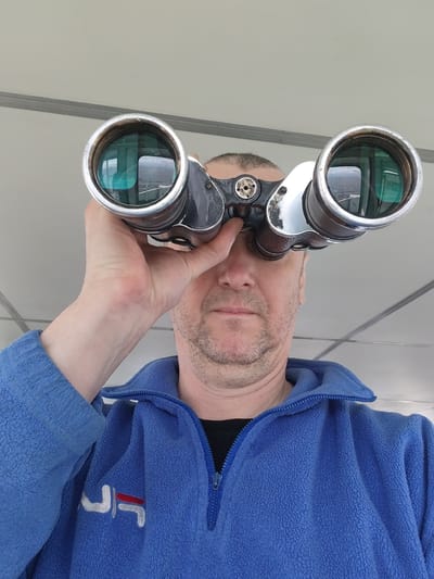 A medium-shot captures a man looking through a pair of binoculars. He is seen from the chest up, wearing a blue zip-up fleece jacket with a white and red logo on the left side. The binoculars are silver and black. The reflection in the binoculars reveals a water view with an exit sign. The background is a blurred white ceiling with a metal trim. The location in the city of Mologa, Russia, cannot be determined.