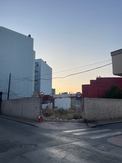 In La Línea De La Concepción, Spain, at dusk, a view of an intersection is captured. A low, concrete block wall runs along the side of the road, separating it from a vacant lot filled with dry grass and debris. Beyond the vacant lot, there are several buildings. On the left, a tall, pale blue apartment building dominates the scene, followed by a shorter, light blue building. To the right of the vacant lot is a bright red building. A speed limit sign for 20 km/h and a pedestrian crossing sign are visible near the red building. A convex mirror is mounted on a pole near the intersection, reflecting the street and buildings. The road surface is asphalt, showing signs of wear and some light reflections from the setting sun. A pedestrian crossing is marked with white stripes. The sky is a gradient of pale blue and orange, indicating either sunrise or sunset.