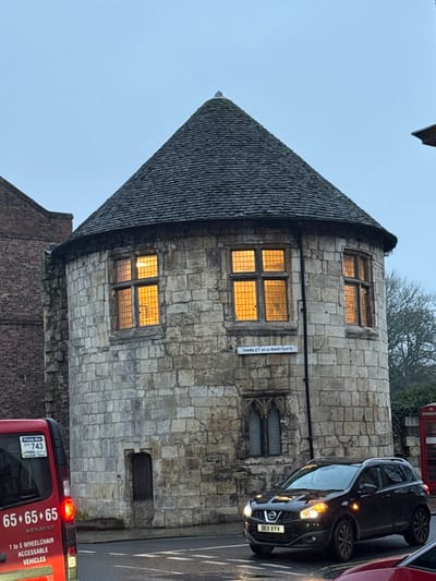 A stone building with a conical roof stands in the foreground, windows glowing from within. A street sign reads "HAMLET of st MARYGATE." To the right, a black car is partially visible, its license plate reading "DEII XYV." Below, a red van displays the number "65.65.65" and text indicating wheelchair accessibility. A "Private" sticker and number "743" are also visible on the van. The sky is overcast, and other buildings and trees are in the background.
