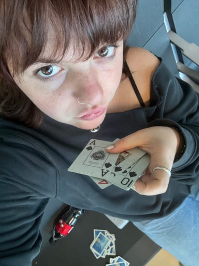 A young woman lies down wearing a dark sweatshirt and jeans. She holds playing cards featuring aces and a ten of spades. On the table next to her are several landscape-printed cards and a miniature Coca-Cola bottle ornament. The woman has brown hair, freckles, and multiple facial piercings. She wears a silver ring on her hand that holds the cards. Her fingernails are painted with glitter polish.
