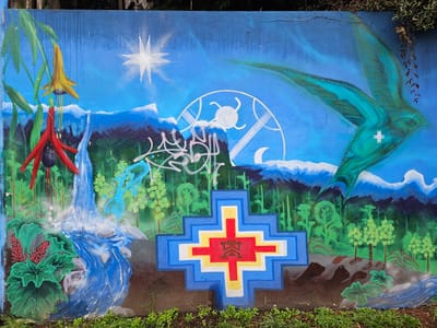 This is a photograph of a vibrant, colorful mural on a wall. The mural depicts a landscape scene with various elements. On the left, there are tropical plants and a waterfall. A bright yellow flower hangs above red, elongated shapes that resemble other flowers or foliage. In the center, a prominent stepped cross, painted in blue, white, yellow, and red, dominates the foreground. Within the central red square of the cross, there is a design resembling two stylized people, or an emblem. Above the cross, there is a depiction of a dense forest with tall, slender trees. A backdrop of blue mountains and sky features a bright white star and a white circular design with radiating lines, a crescent moon is within this design. To the right, a large bird with outstretched wings is painted in shades of green and blue, with a small white star on its body. The overall background is a gradient of blue, suggesting the sky. Graffiti is present on the mural, adding a layer of complexity. The wall is partially obscured at the base by green vegetation and overgrown weeds. The top edge of the wall is visible, with some overhanging foliage. The mural is presumably located in the city of Osorno, Chile.
