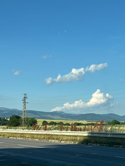 This is a landscape image taken outdoors, likely during the day with clear skies and scattered clouds. The scene depicts a roadside view, with a paved road on the right side of the frame. A metal guardrail separates the road from a grassy embankment. Beyond the embankment, there are rolling hills or mountains covered in vegetation, suggesting a rural or semi-rural location. In the distance, trees and possibly some structures are visible. Power lines stretch across the sky, supported by a tall, lattice-style electricity pylon in the lower right quadrant of the image. The time of day appears to be daytime, with bright sunlight casting shadows. The weather seems pleasant and clear, with a bright blue sky. The image is oriented vertically, which might suggest it was taken from a moving vehicle or held at an angle. There is no visible text in the image. The location context provided is Gorni Bogrov, Bulgaria.