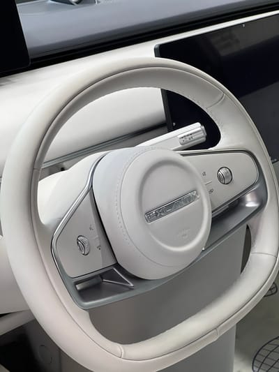 The image shows a close-up view of a white leather steering wheel inside a modern car. The steering wheel has a minimalist design with a central circular hub. The hub features a silver emblem with the text "EXLANTOX" and "AIRBAG" written below it. On the right side of the steering wheel, there are silver buttons and a rotary dial, along with icons suggesting volume control, mute, and mode selection. To the left of the steering wheel, a silver gear selector lever is visible, with indicators for Park (P), Reverse (R), Neutral (N), Drive (D), and possibly manual modes. The dashboard area behind the steering wheel is primarily white and gray, with a black digital display screen on the right. This scene is not located in Kabanikha, Russia; it depicts the interior of a vehicle.