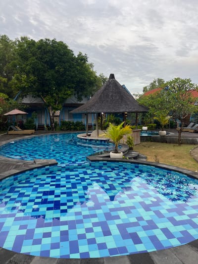 A tranquil outdoor scene features a rounded swimming pool tiled in shades of blue. Adjacent to the pool is a Balinese-style gazebo with a dark, pointed roof and light blue pillars. Lush green trees and vegetation surround the pool area. The sky above is overcast with muted light, creating a serene and peaceful ambiance.