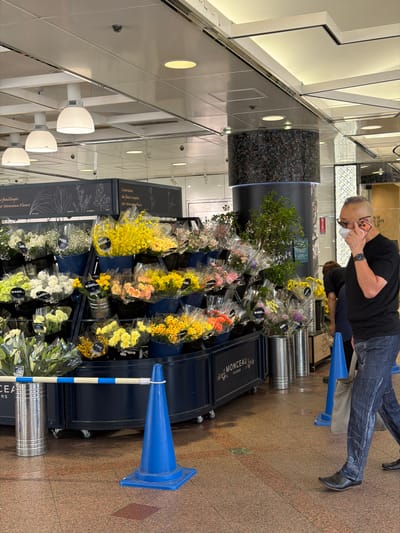 In a commercial setting, a man wearing a black t-shirt, dark patterned jeans, glasses, and a face mask is talking on his phone. He is standing to the right of a flower stall. The stall displays various bouquets of flowers, some wrapped in cellophane and others in buckets. The flowers are in multiple colors, including yellow, white, pink, and orange. Price tags are visible on some of the bouquets. In the background, there is a cylindrical pillar with mosaic tiles and a ceiling with fluorescent lighting. A blue traffic cone is positioned on the floor to the left of the man. The floor is tiled with a reddish-brown pattern. The man is holding a light-colored tote bag in his left hand. The scene appears to be in the city of Chuo, Japan, given the presence of Japanese yen price tags (¥490, ¥300) on the flower bouquets.