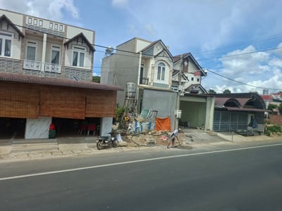 The image depicts a street scene in Phú An, Vietnam, on a cloudy day. In the foreground, a paved road with a white dividing line runs horizontally across the frame. To the right of the road, a man wearing a white shirt and beige pants is engaged in construction work, using a shovel to mix cement on the ground. Adjacent to him is a bucket and construction materials. Further to the right, a building with a grey facade and a closed metal gate is visible.
Behind the construction site, a row of buildings lines the street. The leftmost building is a multi-story structure with a textured facade, ornate balconies with white metal railings, and windows covered by white curtains. To its right, a narrower, multi-story building with a pointed roof features decorative architectural elements and a small balcony. Further down the street, other buildings with pitched roofs and various facade styles are visible, interspersed with trees and vegetation. A parked motorcycle is positioned in front of the first building, near a set of concrete steps leading up to its entrance. A bamboo-like blind covers the lower section of the leftmost building's facade. The overall environment suggests a developing or residential area with ongoing construction activities.