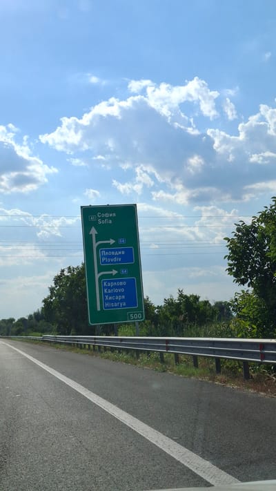 The image shows a green road sign with white text and arrows, indicating directions to various locations. The sign is tilted and appears to be made of metal. The text on the sign is in Bulgarian and English. The top section indicates "A1 София Sofia" with an arrow pointing straight ahead. Below that, there are two blue boxes, each with the number "64" in a white box. The left blue box says "Пловдив Plovdiv" with an arrow to the right. The right blue box says "Карлово Хисарlovо Hisarya" with an arrow to the right. At the bottom of the sign, the number "500" is visible.
The sign is positioned next to a paved road with a white lane marking. A metal guardrail runs along the side of the road. Behind the sign and road, there are trees and greenery, suggesting a rural or suburban setting. The sky is blue with scattered white clouds, indicating daytime. There are power lines visible in the background.
Based on the information on the sign (Sofia, Plovdiv, Karlovo, Hisarya), this road sign is located in Bulgaria. Specifically, it is likely situated on a route leading to these cities. Without further context or identifiable landmarks, pinpointing the exact location in Войводино, Bulgaria, is not possible solely from this image. However, the sign itself is a key element for identifying the road and potential routes within Bulgaria.