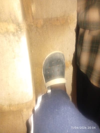 A low-angle, dimly lit, medium shot shows the lower body of a person standing beside a rough, textured, tan-colored wall that appears to be made of concrete or stone. The wall has some faint linear markings and a darker, wet-looking stain near the bottom. To the right of the wall, a person's leg is visible, wearing a dark blue garment, possibly pants or a skirt. On the person's left foot, a black Croc-style shoe with a white sole is worn. The shoe has circular perforations. A portion of another person's torso is visible on the far right, wearing a plaid shirt with dark green and brown patterns. A timestamp, "11/06/2026 20:04," is superimposed on the bottom right of the image. The scene appears to be outdoors, possibly in a street or alleyway within the city of DU, Nigeria, given the presence of the wall and the overall rough texture of the environment.