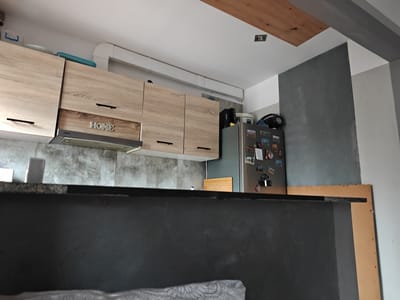This image depicts a modern kitchen in Stargard, Poland. The scene is illuminated by artificial light, suggesting it could be any time of day. The kitchen features light wood-grain cabinets with black handles mounted above a gray tiled backsplash. A stainless steel range hood is installed under the cabinets. A decorative metal sign with the word "HOME" is centered on one of the cabinet doors.
To the right of the cabinets, a stainless steel refrigerator is visible, adorned with various magnets and photos. Above the refrigerator, there's a portion of a dark gray wall. The ceiling is white, with a wooden plank accent and a recessed light fixture. A white pipe runs along the ceiling. In the foreground, a dark gray countertop or partition is visible, with a rough texture. Part of a light-colored fabric with a patterned design is seen at the very bottom of the image. The overall aesthetic is contemporary and functional.