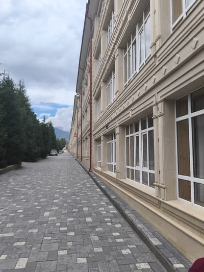 The image shows a pathway in front of a modern beige building with numerous white-framed windows. The pathway is paved with interlocking gray and white rectangular stones. To the left of the pathway are tall, dark green evergreen trees. In the distance, there are cars parked along the pathway and mountains are visible under a cloudy sky. A red sign with white text is visible on the building in the distance. The building's facade is decorated with horizontal lines and square indentations. A narrow drainage channel runs along the base of the building. There is no visible indication of the city of Bayet, Kyrgyzstan in the image.