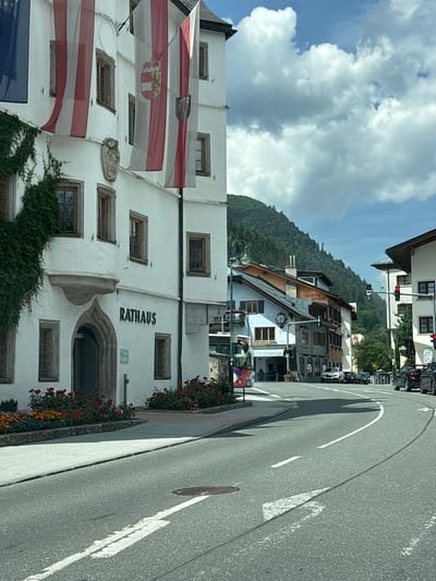 A white building with a "RATHAUS" sign in black, which signifies town hall, is the main subject. The building is decorated with Austrian flags and has ivy growing on its side. In front of the building, there are flower beds with red and orange flowers. The street in front of the building is paved and has white lane markings. Several cars are parked along the street and on the side. In the background, there are more buildings and a lush green mountain covered in trees. The sky is blue with white clouds. The scene appears to be in a town in Austria, specifically Zell am See, as suggested by the presence of Austrian flags and the architecture.