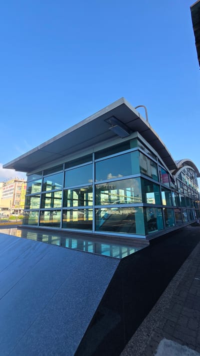 The image shows the exterior of a modern building with large glass windows. The building is made of glass and steel, with a flat roof and a curved section on the right side. The glass windows reflect the sky and the surrounding buildings, which appear to be a mix of residential and commercial structures. The building is located on a street with a sidewalk and a paved road. The sky is clear and blue, with a few clouds visible. The overall scene suggests a sunny day in an urban environment. The building is the **Kaohsiung Exhibition Center (KEC)**.