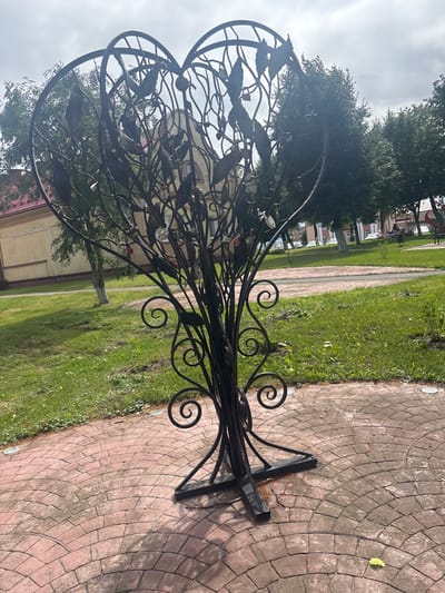 This media file shows a black metal sculpture of a tree with ornate swirling branches and leaf-like decorations. The sculpture is positioned on a red-tiled pathway with grass and trees in the background. The scene appears to be outdoors in a park or public space, possibly in the Капыльскі сельскі Савет, Belarus, given the provided location context. The sky is overcast, suggesting a cloudy or possibly rainy day, and the lighting is diffused. There is no visible text in the image. The sculpture itself is the primary subject, and it is intricately crafted, giving it an artistic and decorative appearance. The overall atmosphere is calm and serene, with the sculpture serving as a focal point in the landscape.