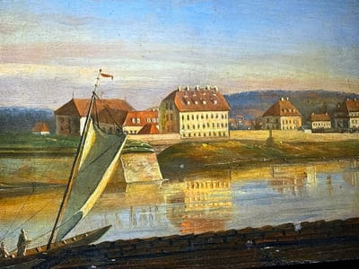 This detailed painting captures a serene riverside cityscape, purportedly depicting a location within the city of Reus, Spain. The artwork features visible brushstrokes, indicative of an oil painting.

In the immediate foreground, a dark, textured strip, possibly representing a dock or the near bank of the waterway, runs along the bottom edge. Above this, a broad body of water, likely a river or large canal, occupies the lower and central portions of the scene. The water surface reflects the sky and the distant buildings, displaying a mix of golden, warm hues and cooler blues, with subtle ripples distorting the reflections.

On the left side of the water, closer to the viewer, a wooden sailing boat is positioned. It has a tall, slender mast that leans slightly to the right, from which a large, light-colored (possibly off-white or yellowish) sail is partially unfurled or draped. A small red pennant or flag flies distinctly from the masthead. One figure, dressed in muted tones, is visible standing near the stern of the boat, appearing to be attending to the vessel.

Across the water, the far bank is populated by a cluster of architectural structures. A prominent multi-story building with a light-colored facade and a distinctive red-tiled hip roof, featuring several white chimneys or dormer windows, stands centrally. It exhibits numerous evenly spaced windows. To its left, another building with a red roof and what appears to be a square or tower-like section is visible. Further to the right of the central building, additional structures with similar red roofs and light-toned walls extend along the bank. A light-colored stone structure, possibly a bridge pier or embankment, projects into the water from the far bank, casting a clear reflection.

In the distant background, rolling hills or low mountains are depicted in soft, muted tones of green and brown, suggesting a natural landscape beyond the urban area. The sky above is a clear light blue, transitioning to warmer, lighter hues of pale yellow and orange near the horizon, indicative of either an early morning or late afternoon light illuminating the scene with a soft, golden glow that highlights the buildings and water. The overall atmosphere is tranquil, with the boat serving as the primary point of human activity within the calm environment.