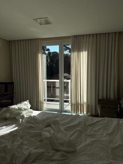 The image displays the interior of a room, located in Canela, Brazil. A large bed in the foreground is covered with disheveled white sheets and a duvet. Strong natural light enters through a tall, white-framed glass double door, positioned centrally in the background, which leads to an outdoor tiled balcony. Floor-to-ceiling light-colored curtains are partially drawn back on either side of the glass door. To the left of the bed, a dark wooden chair is partially visible. To the right, a light-colored armchair with a dark bottle on an adjacent surface is present. Beyond the balcony, a red-brick building with a brown roof is visible, partially obscured by dense, dark green trees. A clear blue sky is visible above the trees. The room has a light-colored ceiling with a square, recessed light fixture. No individuals are present in the scene.