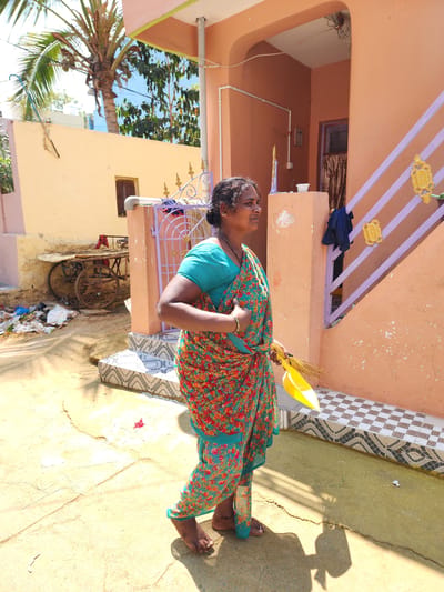 An adult woman is depicted in an outdoor residential area of Pedda Kammavaripalli, India, under clear, sunny conditions. The woman, barefoot, is shown in profile, facing right. She has dark skin, dark hair tied back, and wears a teal-colored blouse beneath a vibrant floral sari with red, orange, green, and teal patterns. She is adorned with a gold-colored bangle on her right wrist and a dark beaded necklace, with a small stud in her right nostril. Her left hand holds a yellow plastic dustpan containing a natural fiber broom.
The immediate ground is a cracked, light-colored concrete or paved surface, exhibiting distinct shadows. To the woman's right, a peach-colored house features a tiled base with a black and white geometric pattern, and purple stairs/railing with two ornate golden embellishments. A dark blue garment hangs over a structural pillar of the house. An arched entrance leads into a darker interior with a brown curtain, and exposed electrical wiring is visible overhead.
In the midground to the left, another faded peach-colored building has a small, dark window. Adjacent to this building, a pile of assorted refuse (plastic, paper) accumulates next to a rustic, two-wheeled cart. Above this structure, a large palm tree with visible coconuts stands against a bright blue sky. White PVC piping runs horizontally on the walls of both houses.