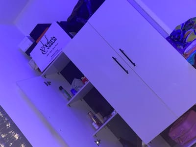 A white cabinet with black handles stands against a wall, one door slightly ajar revealing interior shelves. The shelves hold items like bottles and containers, partially obscured in shadows. Atop the cabinet, boxes and a shopping bag are stacked, the bag bearing the logo "Chris Vogue." A patterned garment rests partially on the cabinet's upper right. The scene is illuminated with a distinct blueish-purple cast.