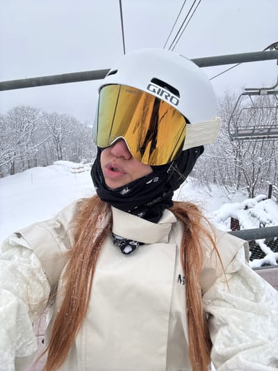 A person is on a ski lift, wearing a white helmet with the word "GIRO" on it, and gold-tinted goggles. They have a black neck warmer and a light beige jacket. The person has long reddish-brown hair that is covered in snow. In the background, there are snow-covered trees and ski lifts. The weather appears to be snowy.
