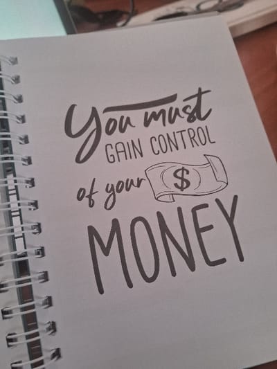 This image, taken in Muthambi ward, Kenya, displays a white spiral-bound notebook open to a page featuring motivational text and a graphic. The text reads: "You must GAIN CONTROL of your MONEY." The phrase "You must" is rendered in a script font, "GAIN CONTROL" is in uppercase sans-serif, "of your" is in a script font, and "MONEY" is in large, bold, uppercase sans-serif. Adjacent to "of your" and above "MONEY" is a simple outline drawing of a rolled banknote with a dollar sign ($) symbol. The notebook's white plastic spiral binding is visible along the left edge of the page. The background appears blurred, suggesting an indoor environment, possibly a desk or table, with indistinct objects partially visible in the upper right quadrant. No individuals or explicit actions are depicted.