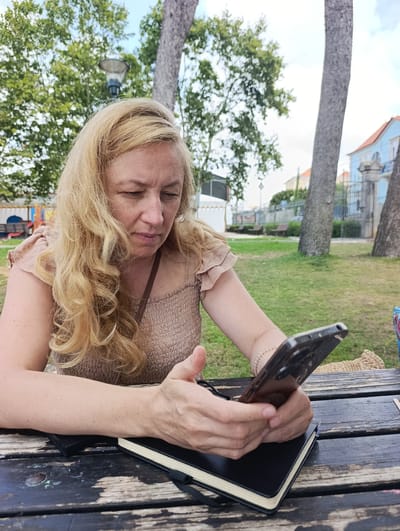 A blonde woman is sitting at a rustic wooden picnic table in a park. She is wearing a light brown, off-the-shoulder top. The woman is looking down at her phone, which she is holding in her hands. Her left hand is resting on a black notebook placed on the table. The park has green grass, trees, and a lamppost in the background. In the distance, there are buildings, a fence, and benches. The scene appears to be in Parede, Portugal.