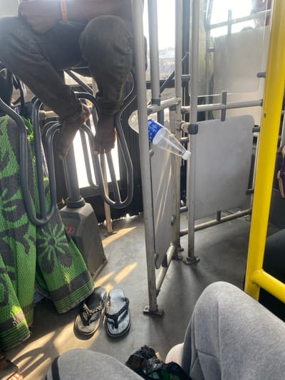 The image depicts the interior of a public transport vehicle in Igando Oloja, Nigeria, during daytime, characterized by strong natural light casting shadows on the floor. In the upper left, an individual is partially visible from the waist down, wearing dark trousers and resting bare feet on a series of horizontal metal support bars. A green and dark patterned blanket or fabric is draped over adjacent vertical metal bars. On the floor, beneath the person's feet, are two pairs of black flip-flops. To the right, a clear plastic water bottle with a blue label is wedged horizontally between two metal bars. Further right and in the foreground, the lower legs of another individual, wearing grey sweatpants, are visible, suggesting a seated position. A crumpled black plastic bag rests near their feet. The vehicle's interior features multiple grey metal poles and panels creating passenger partitions and handholds.