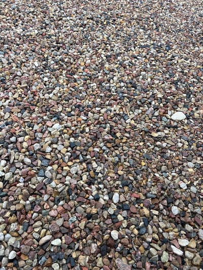 The image shows a ground-level view of a large area covered in gravel. The gravel consists of small, irregularly shaped stones of various colors, including shades of grey, brown, red, pink, and white. Some stones appear wet, suggesting recent rain or a damp environment. There are no people or discernable objects other than the gravel itself. The scene depicts an outdoor ground surface. The location in Капыльскі сельскі Савет, Belarus, cannot be determined from the image alone.