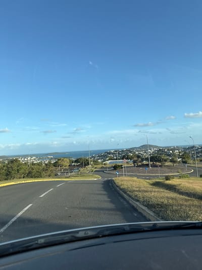 The image depicts an elevated, daytime view from within a vehicle, with the top of the dashboard visible at the bottom of the frame, looking down a road towards a coastal city and bay in Nouméa, New Caledonia, an overseas territory of France.
The foreground features an asphalt road, marked with solid and dashed white lines, which descends and curves towards a multi-lane roundabout. To the left of the initial road, green foliage is visible. To the right, a strip of dry, yellow-brown grass separated by a concrete curb borders the road.
The midground is dominated by the circular roundabout, which has a central island covered with sparse, dry vegetation and several small trees. Multiple blue and white traffic signs, including a yield sign and a roundabout directional sign, are visible on the perimeter of the central island. Several streetlights with curved arms are positioned around the roundabout. Beyond the roundabout, to the left, additional road segments and a small parking area with parked vehicles are discernible.
The background reveals a sprawling urban landscape of predominantly light-colored buildings spread across green, undulating hills. Beyond the cityscape, a large, blue body of water, characteristic of a bay or ocean, stretches towards the horizon. Several landmasses or islands are visible within the water. The sky above is clear blue with scattered, wispy white clouds. No individuals or actively moving vehicles are present in the immediate foreground or midground.