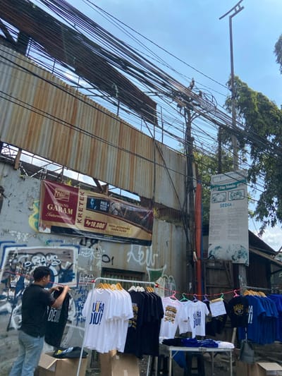 The scene is a street corner in Kecamatan Cimahi Selatan, Indonesia, at the coordinates -6.909 latitude and 107.568 longitude. A man in a black shirt and blue jeans stands in front of a corrugated metal fence, looking at something just out of frame. He is holding a black garment in his right hand. The fence is covered in graffiti and has several wires running across it. A sign advertising "Gudang Garam International" kretek cigarettes is on the wall to the left, with a price of Rp 18.000 for the cigarettes. To the right, a sign in Indonesian advertises "Sedia" - meaning "available" - with a list of items. A vendor's stand is set up in front of the wall, displaying rows of shirts on a metal rack, along with boxes and other merchandise. There are various colors of shirts, with some featuring logos and text, including "URA DJU!" and "Persib". The background is a bright blue sky, with several green trees visible in the distance. Many electrical wires run across the sky and down the side of the building. The scene appears to be located in a densely populated, urban area.