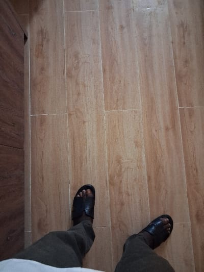 The image depicts an overhead view of an individual's lower legs and feet in an indoor environment located in the city of Takum, Nigeria. The person is wearing dark-colored, open-toed sandals and olive-green trousers. They are positioned centrally on a floor composed of rectangular light brown tiles featuring a wood-grain pattern, with visible lighter-colored grout lines. A dark brown, vertical wooden surface or furniture component is present along the left edge of the frame.