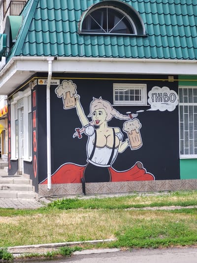This is a photograph of a mural on the side of a building, likely a business, in Zolotonosha, Ukraine. The mural depicts a stylized cartoon woman with blonde pigtails, wearing a traditional Bavarian-style outfit including a white blouse and black suspenders, and holding two large mugs of frothy beer. A speech bubble above her head contains the word "ПИВО" (Pivo), which is Russian for beer. The building has a distinctive green, wavy, tiled roof and white gutters. To the left of the mural, there are white shuttered windows and a visible street sign with Cyrillic text. The building is situated next to a paved walkway and a grassy area, with a street visible in the foreground. The overall scene suggests a commercial establishment, possibly a pub or bar, that uses vibrant artwork to attract customers.