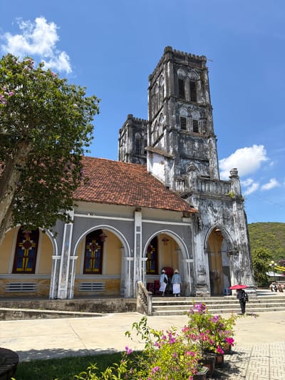 The image depicts the exterior of a stone church in Hội Tín, Vietnam, under a clear blue sky with scattered clouds. The church features a central section with a red tile roof, flanked by two weathered stone towers. Arched windows with stained glass are visible on the side of the main building.

In the foreground, a lush green tree with pink blossoms stands to the left, and pots of vibrant pink and purple flowers are arranged in front of the church entrance. Several individuals are present. Two people wearing white robes and hats stand near the entrance of the church. Another person, holding a red umbrella, walks away from the camera towards the steps of the church. In the background, more people are seated on a bench, and string lights are visible. The landscape behind the church is characterized by green, rolling hills. The overall scene suggests a bright, sunny day at a historic religious site.