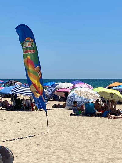 The image depicts a crowded beach in Conil de la Frontera, Spain, on a clear, sunny day. In the foreground, a blue flag with a rainbow-colored popsicle graphic and the word "TROPICAL" written on it waves from a pole. The beach is filled with numerous beach umbrellas and chairs, providing shade for beachgoers. Several people are visible, either sitting under umbrellas, lying on towels, or walking on the sand. The ocean is visible in the background, with a clear horizon. The overall scene conveys a typical summer day at a popular beach destination.