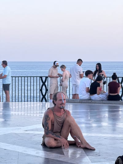 In the foreground, a man with facial paint and arm tattoos is sitting cross-legged on a patterned stone floor, looking to the left. He is wearing a tan garment. In the background, several people are standing and sitting on a ledge overlooking a body of water. The scene appears to be a promenade or viewing area in Nerja, Spain, with the Mediterranean Sea visible beyond a black metal railing. The sky is overcast and pale blue.