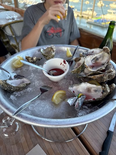 A person with a glass is visible to the left of a platter of oysters on ice. A green bottle is on the side of the platter, alongside a knife. Oyster forks are scattered, and a small cup of sauce is visible with a spoon. Lemon wedges are among the oysters. The scene is on a wooden table, with a wine glass slightly out of focus on the right.