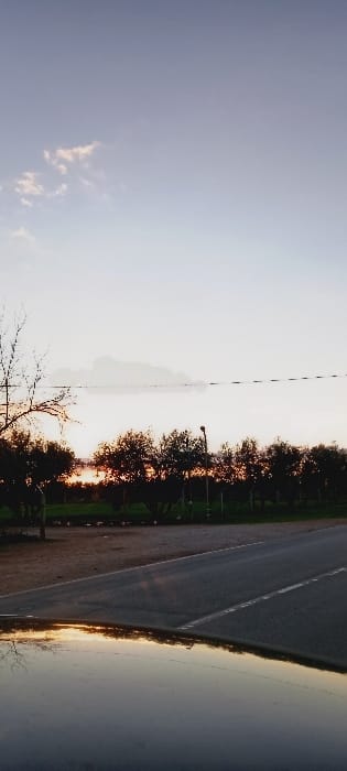 This image, set in Dar Oum Soltane, Morocco, captures a tranquil landscape scene during either sunrise or sunset. The sky, occupying the upper portion of the frame, transitions from a soft blue-grey to a bright, almost white hue near the horizon, indicating the low position of the sun. A small, faint, wispy cloud is visible in the upper left corner.

A prominent line of trees stretches horizontally across the middle of the image, their dark forms silhouetted against the glowing light of the sun behind them. This backlighting creates a warm, orange-yellow glow around the foliage. A dark silhouette of a streetlight can be discerned among the trees.

In the foreground, an asphalt road runs parallel to the treeline, visible on the left side of the frame with a white lane marking and a dirt shoulder. To the right of the road, a calm body of water, possibly a river or lake, reflects the bright sky and the warm light, creating a shimmering effect. Bare tree branches extend into the lower corners of the image.

No people or discernible activities are present, emphasizing the serene and natural beauty of the scene. The warm lighting and long shadows characteristic of the golden hour evoke a sense of peace and stillness. There is no visible text in the image.