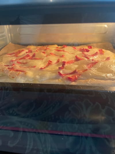 The image shows a close-up view of a pastry with pink toppings baking inside an oven. The pastry is elongated and appears to be covered in a thin, translucent dough, with strips of pink food scattered across its surface. The pastry rests on a piece of parchment paper, which in turn sits on an oven rack. The oven interior is visible, with metallic surfaces and light reflecting from the heating elements. The oven door has a dark, tinted glass, through which the pastry is viewed. On the glass, there are faint, swirling patterns, possibly condensation or residue. The overall lighting is warm, suggesting the oven is in use. The setting appears to be a kitchen. There is no visible text in the image. The context of Bujum, Kyrgyzstan is noted.