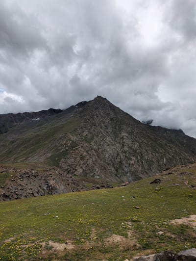 The image depicts a mountainous landscape in Patseo, India, under a cloudy sky. A large, rugged mountain dominates the background, with rocky slopes and patches of greenery. The foreground features a grassy meadow dotted with small yellow flowers. The overall scene is natural and serene, with no visible people or man-made structures apart from a possible fence line in the mid-ground. The lighting suggests it is daytime, but the cloud cover creates a somewhat muted atmosphere.