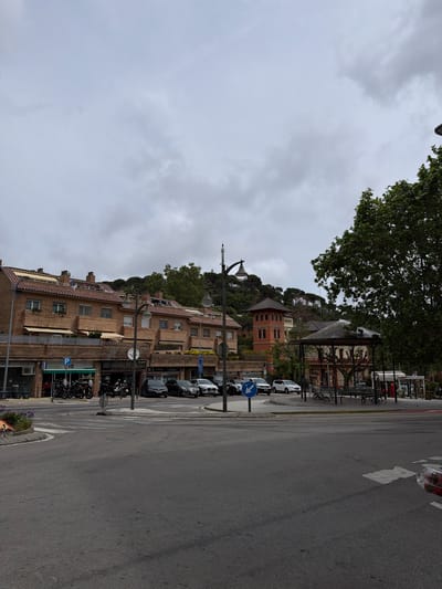 This is an outdoor scene in Sant Andreu de Llavaneres, Spain. A street lined with brick buildings on one side is visible. These buildings have red-tiled roofs, windows, and some have awnings. Several parked cars and motorcycles are along the street. Street signs and lamp posts are also present. The road surface appears to be asphalt. In the background, a building on a hill can be seen amidst trees. The sky is overcast.
