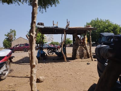 The image depicts a scene in Tongo, Nigeria, located at latitude 10.664 and longitude 11.364. A small, wooden, open-sided structure with a corrugated metal roof, built from branches and timber, stands in the middle ground, with a sign visible in the background that reads "WEDDING, PLEASANT, SMILING, WELCOME." There are two men seated on the ground, facing each other, to the left of the structure. A motorcycle is parked near the structure. A red car is visible parked in the background, and a black car is visible parked to the right of the structure. A tall tree with bare branches is located on the left side of the frame, with a large brown branch that takes up most of the image. The ground is bare, dirt, and dry. The image is likely taken during the day, and the sky is clear.