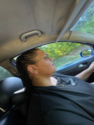 A medium shot captures a woman driving a car on a sunny day, with a blurred green and yellow landscape passing by outside the window. She is wearing a black t-shirt with a cross necklace and glasses. The interior of the car is visible, including the ceiling handle, dashboard, and a portion of the passenger seat. The image appears to be taken from the back seat, looking forward.