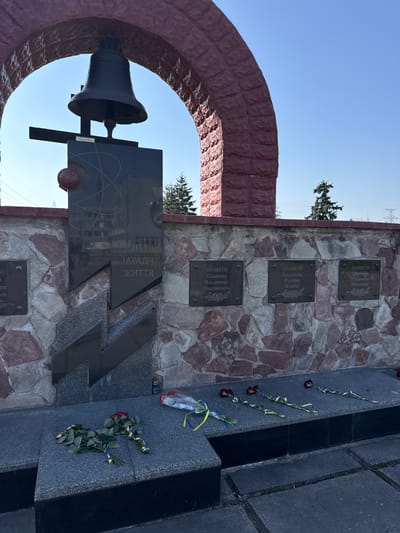 This image depicts a memorial in Pryp'yat', Ukraine, dedicated to the victims of the Chernobyl disaster. The memorial features a bell encased within a red brick arch, and a dark granite monument engraved with an image of a nuclear symbol, and the words "ЗАРАДИ ЖИТТЯ" (FOR LIFE) in Ukrainian. Adjacent to the monument is a wall of stone containing bronze plaques with names of the deceased. Flowers, including roses, and ribbons in the colors of the Ukrainian flag lie at the base of the memorial. Power lines are visible in the background.
